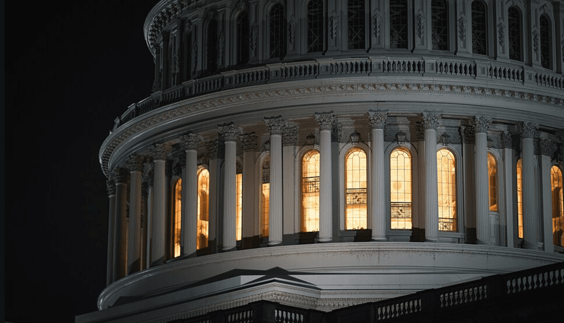 Fotografía nocturna en primer plano de la cúpula del Capitolio de EE. UU. Las columnas de mármol blanco y la mampostería de la cúpula están iluminadas de forma espectacular desde el interior, con una cálida luz dorada que resplandece a través de las altas ventanas arqueadas que rodean la rotonda central, mientras que el cielo circundante es completamente negro.